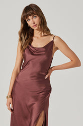 A woman is wearing a sleeveless, satin slip dress in a deep mauve or dusty rose color. The dress has thin spaghetti straps and a cowl neckline. It features a side slit that reaches above the knee. The fabric has a smooth and shiny texture, giving it an elegant appearance.
